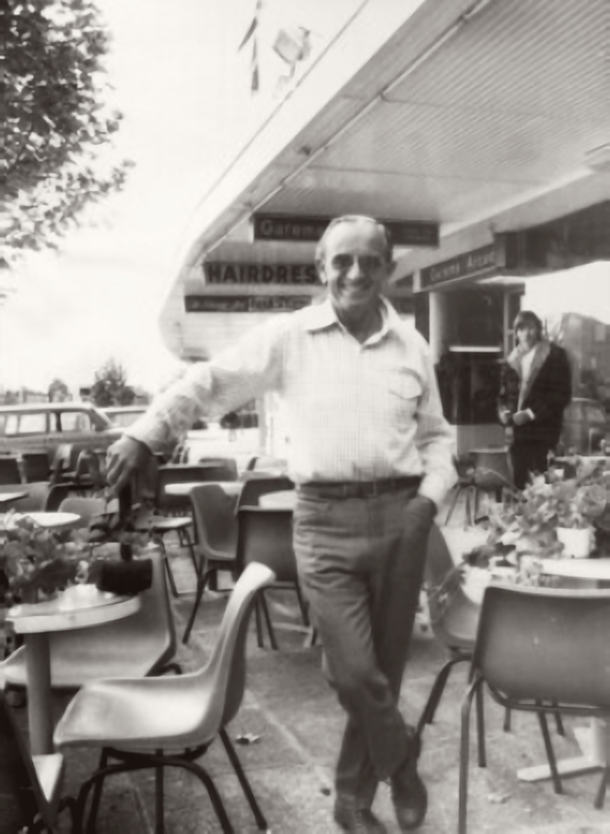 Black-and-white photograph of Augustin (Gus) Petersilka leaning on a chair among the outdoor tables of his café, with the arcade awning behind him.