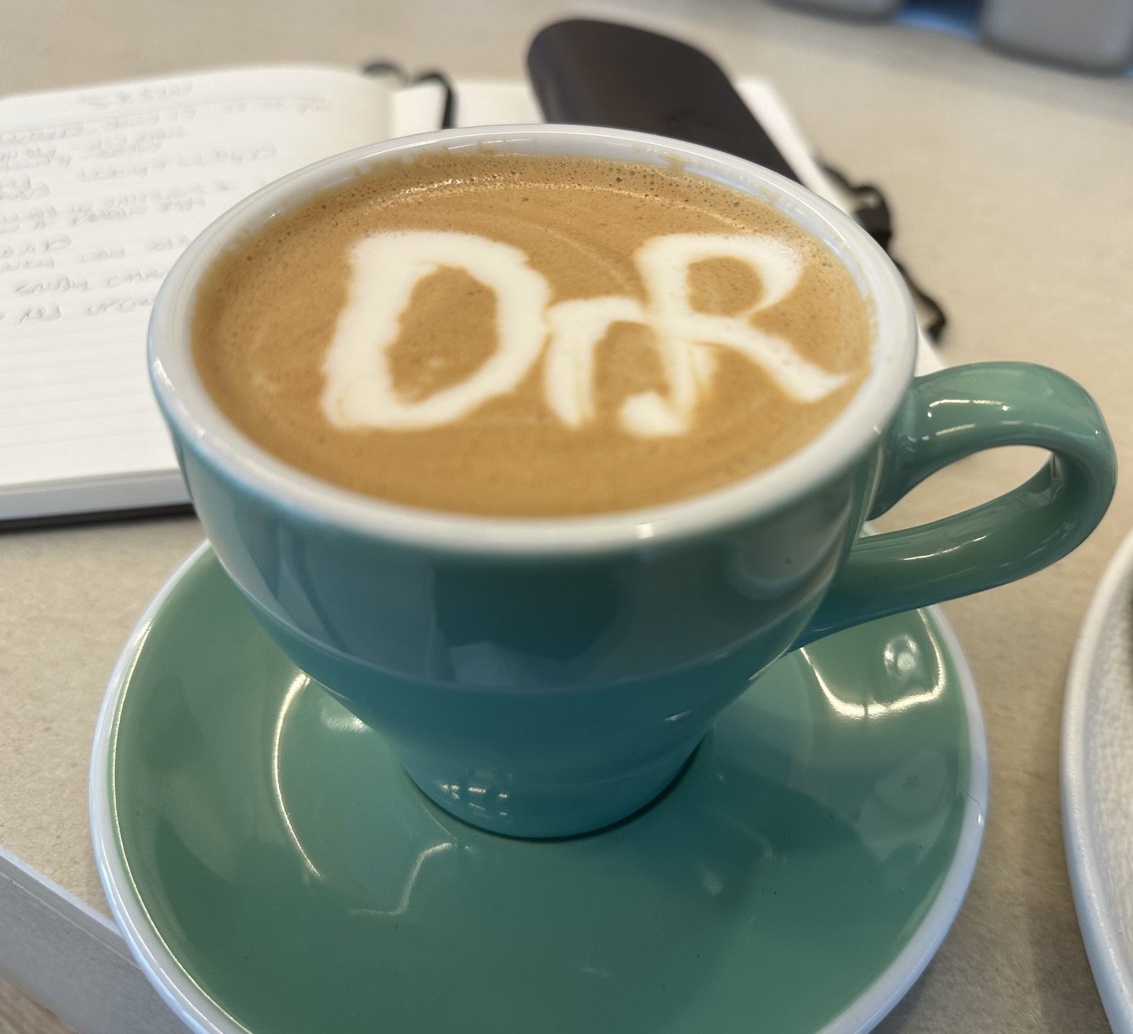 A flat white in a duck-egg green cup, the initials Dr R drawn into the foam by the co-owner, beside an open notebook of handwritten fieldnotes.